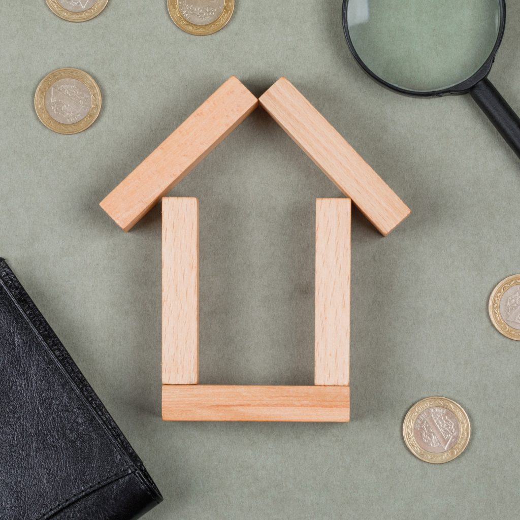 Maqueta de una casa construida con bloques de madera, rodeada de monedas y una lupa, simbolizando la búsqueda de financiamiento hipotecario completo.